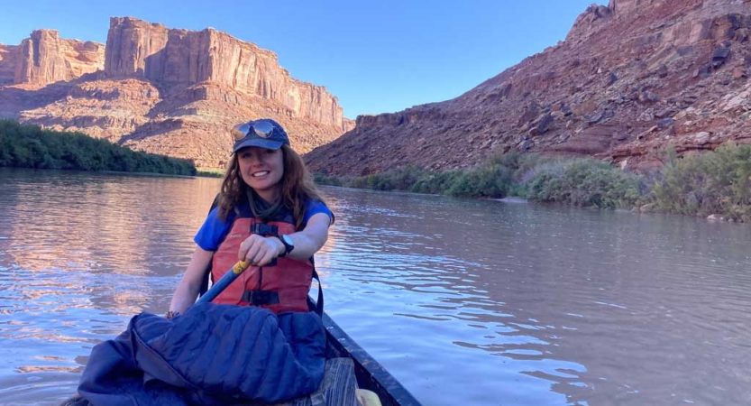 A person smiles while paddling a canoe along a river framed by red canyon walls. 